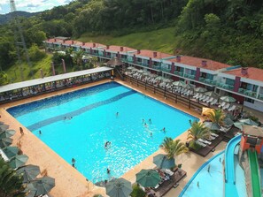 En stor swimmingpool med flere baner, omgivet af liggestole og parasoller, og en bygning med balkoner i baggrunden.