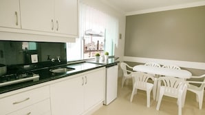 A kitchen with white cabinets, a black countertop, a sink, and a small refrigerator. There is a white table with four chairs and a window with white curtains. - Villa Di Acqua Hotel (Gaspar)