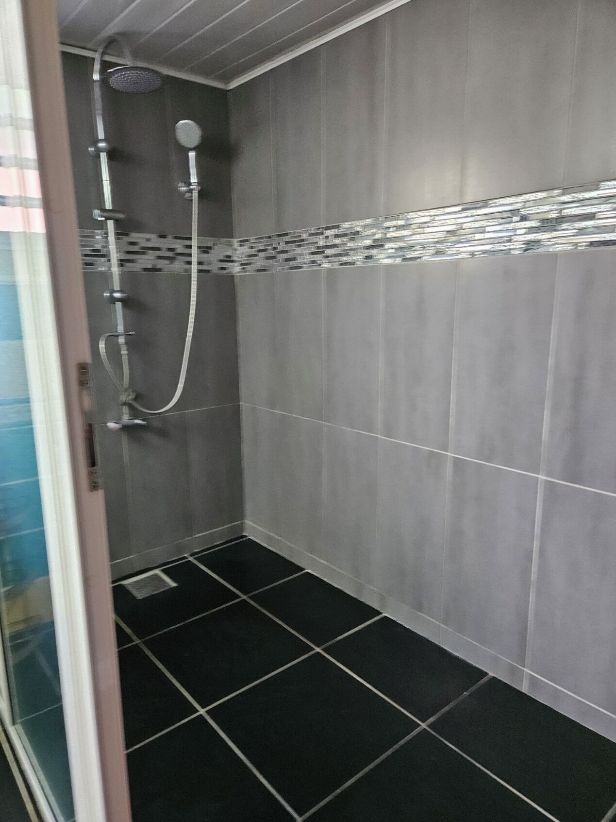 Ducha con puerta de vidrio, paredes de azulejos grises y suelo de losetas negras.
