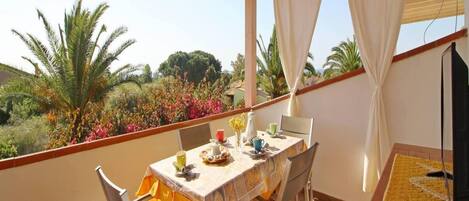 Un espacio comedor al aire libre con techo, una mesa dispuesta para dos personas, sillas alrededor y vista a un jardín con palmeras y flores de colores.