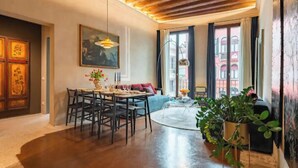 Ein Essbereich mit einem runden Tisch, Stühlen und einem Blumenstrauß. Ein Sofa und ein Couchtisch mit einer Pflanze sind ebenfalls vorhanden. Der Raum verfügt über Holzbalken an der Decke und große Fenster.