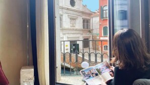 Eine Person liest eine Zeitschrift auf einem Balkon mit Blick auf ein historisches Gebäude.