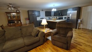 A living room with a sofa, armchair, and a coffee table. There is a kitchen in the background with cabinets and appliances.