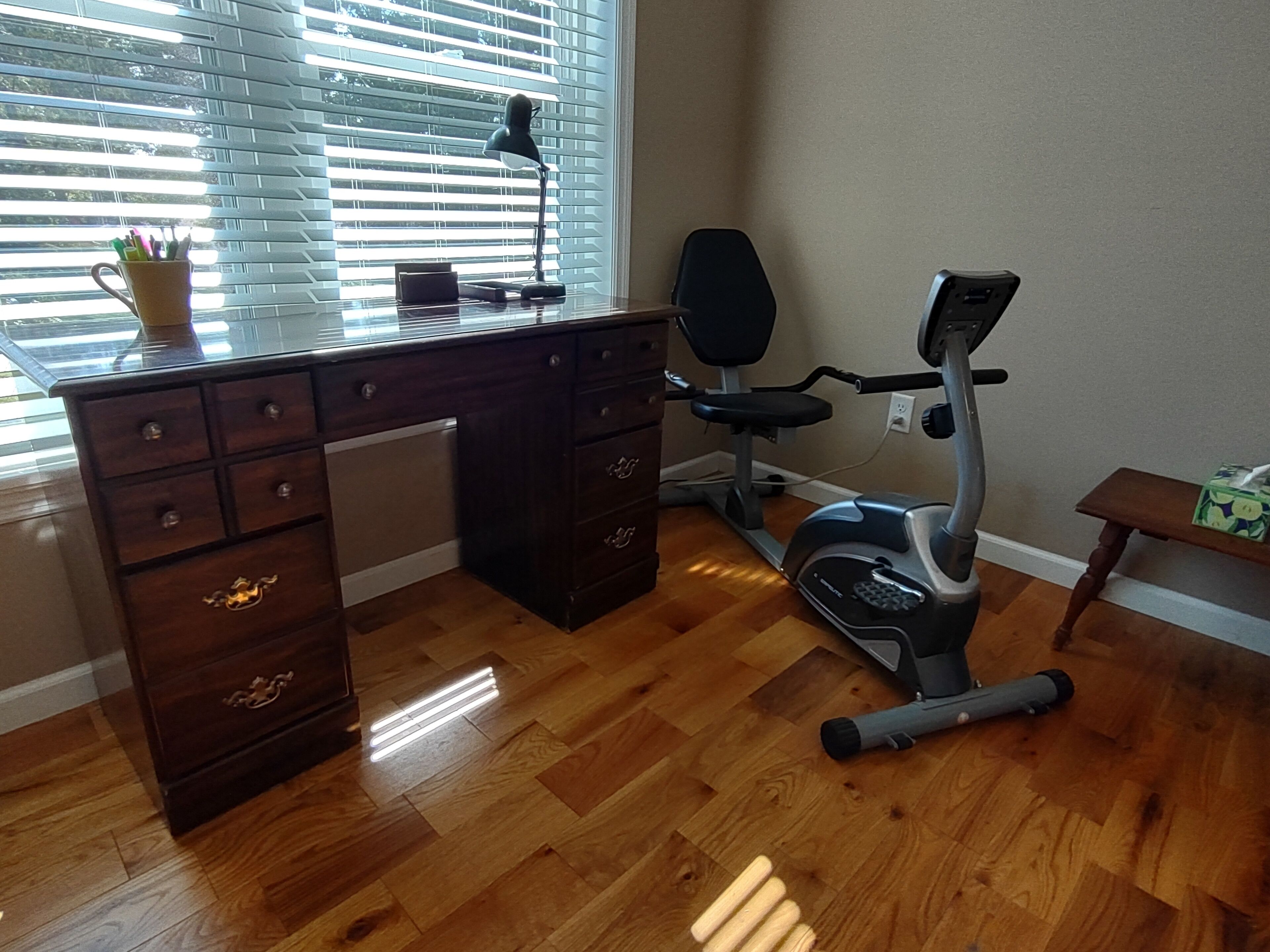 Una habitación con un escritorio de madera, una bicicleta estática y una ventana con persianas.