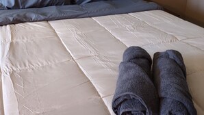 A neatly made bed with a white mattress, grey pillows, and two rolled grey towels.