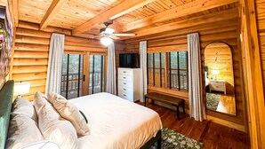 A wooden cabin bedroom with a bed, curtains, a bench, a TV, and a ceiling fan. - Spacious Cabin for 14 on Fightingtown Creek – Private Trout Fishing Retreat (McCaysville)
