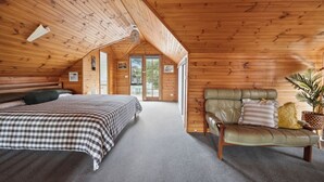 Une chambre mansardée chaleureuse, avec une couvre-lit à carreaux, un canapé en cuir agrémenté de coussins et une vue sur l’extérieur grâce à une porte vitrée.