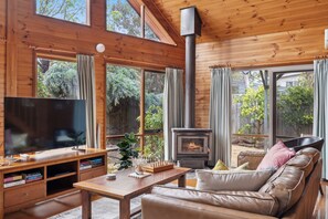 Ein gemütliches Wohnzimmer mit großem Fernseher, Holzmöbeln, einem Kamin und großen Fenstern mit Blick auf einen Garten.