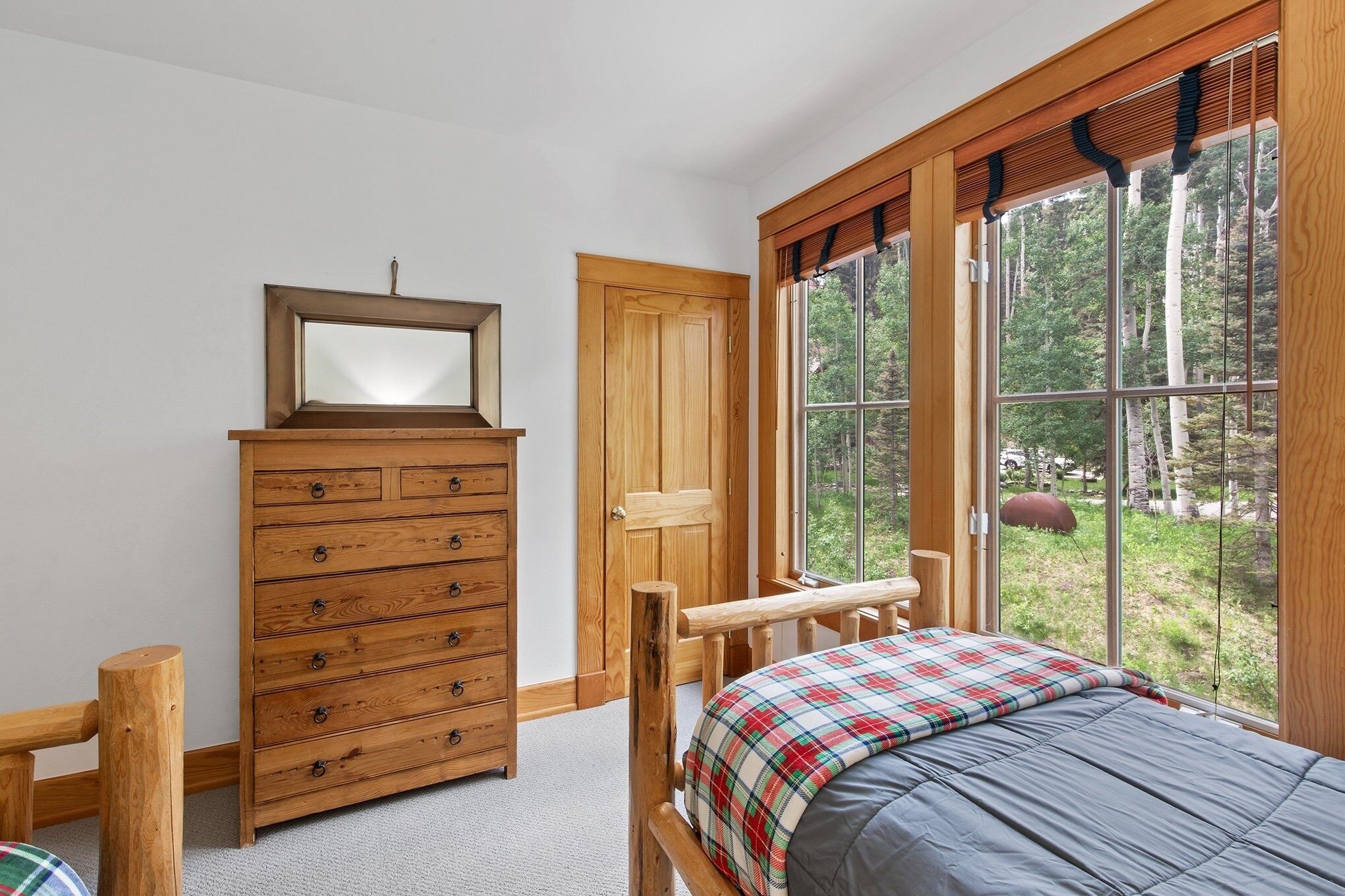Une chambre avec une commode en bois, un lit recouvert d’une couverture à carreaux, une fenêtre donnant sur des arbres et un banc en bois.