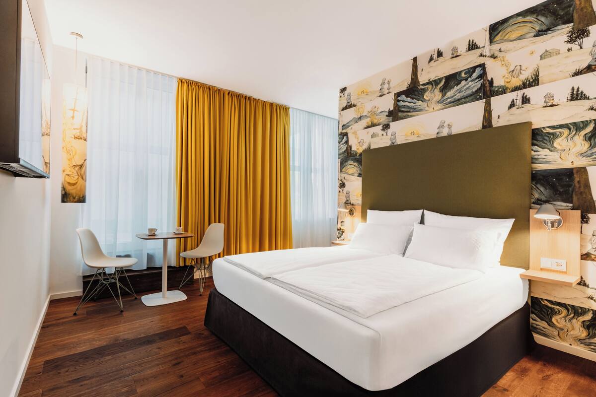 Une chambre d’hôtel avec un grand lit, une petite table, deux chaises et un mur orné d’une fresque à motifs.