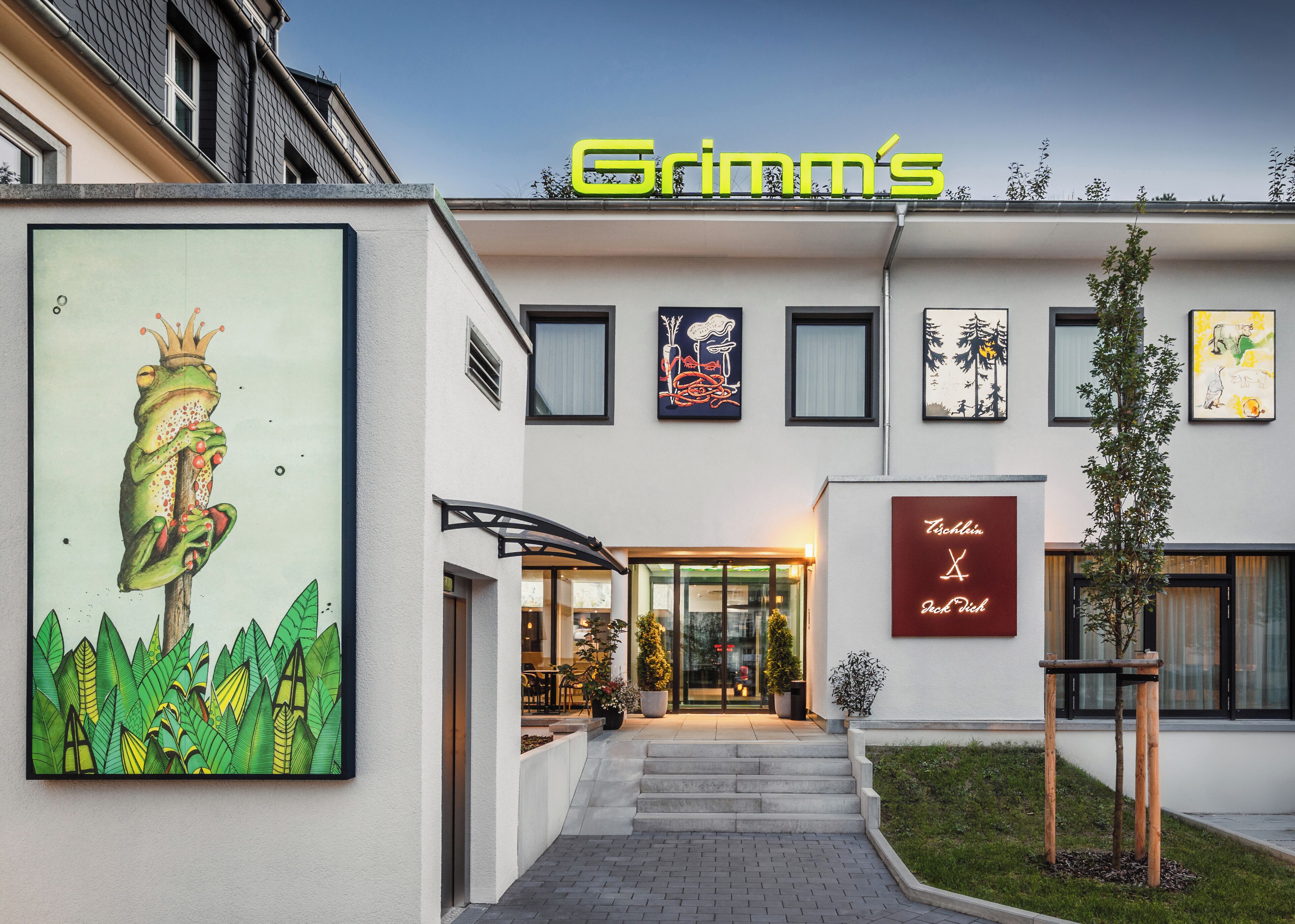 Un edificio moderno con un letrero que dice "Grimm's" en la parte superior. A la izquierda, hay una obra de arte enmarcadas de una rana en una rama de árbol. La entrada tiene una puerta de cristal y un conjunto de escalones que conducen a ella.