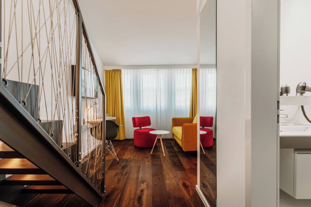 Un salon moderne avec un escalier, un plancher en bois, des fauteuils rouges et jaunes, et une grande fenêtre avec des rideaux.