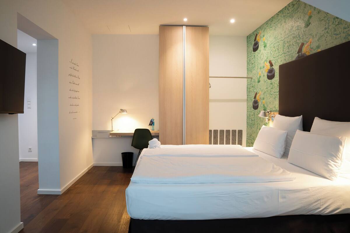 Une chambre d’hôtel moderne avec un grand lit, un bureau avec une lampe, une armoire et un mur orné d’un motif vert.