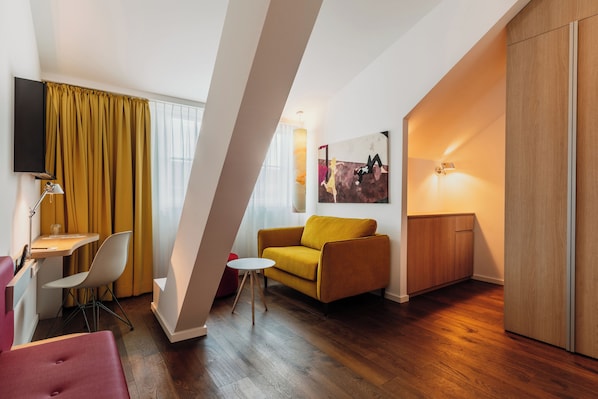 A modern living room with a sofa, a small table, and a chair. There is a TV mounted on the wall and a lamp on a side table. - Grimms Hotel Hanau (Hanau)