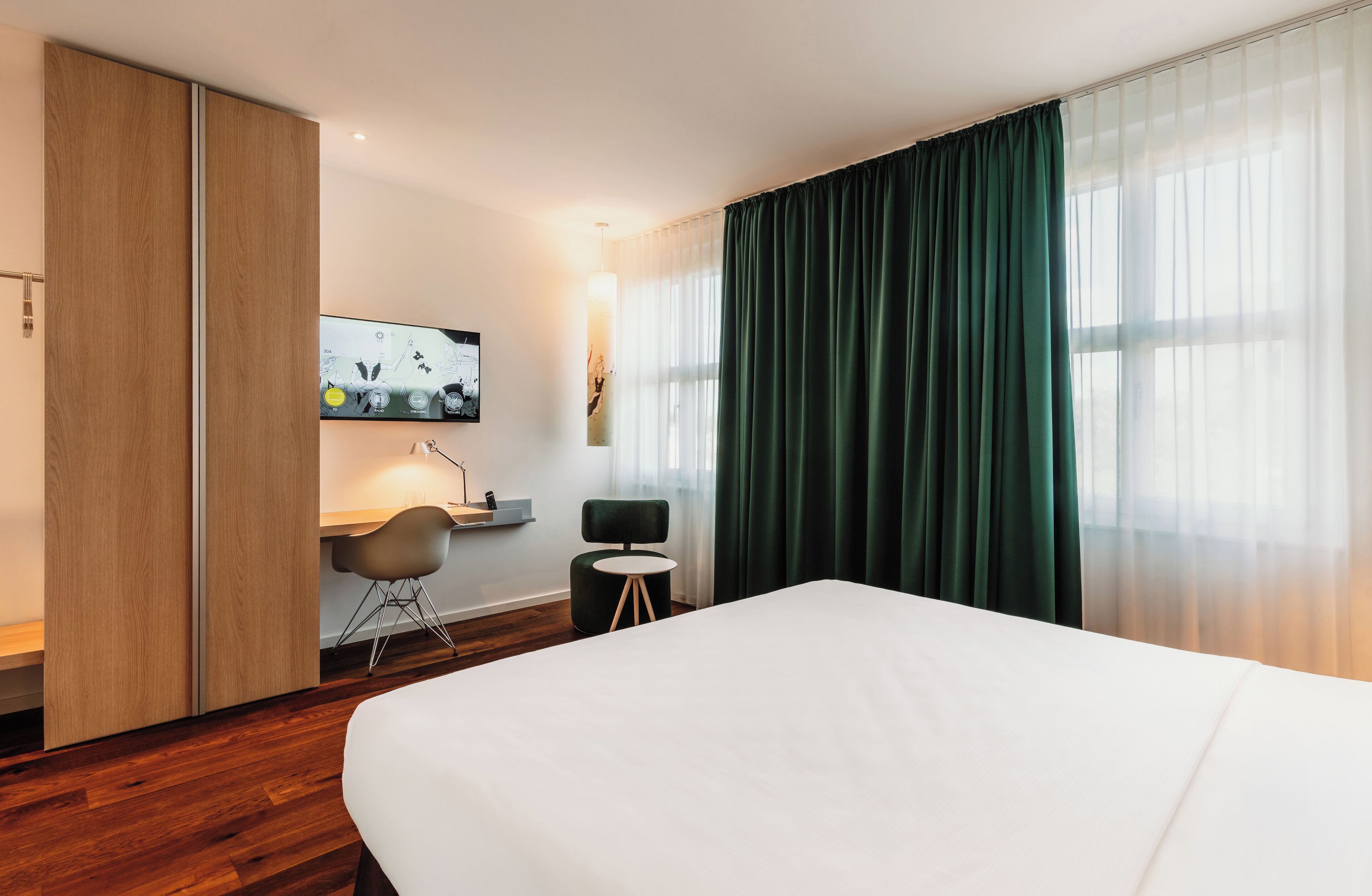 A hotel room with a large bed, a desk with a chair, a TV mounted on the wall, and a window with green curtains.