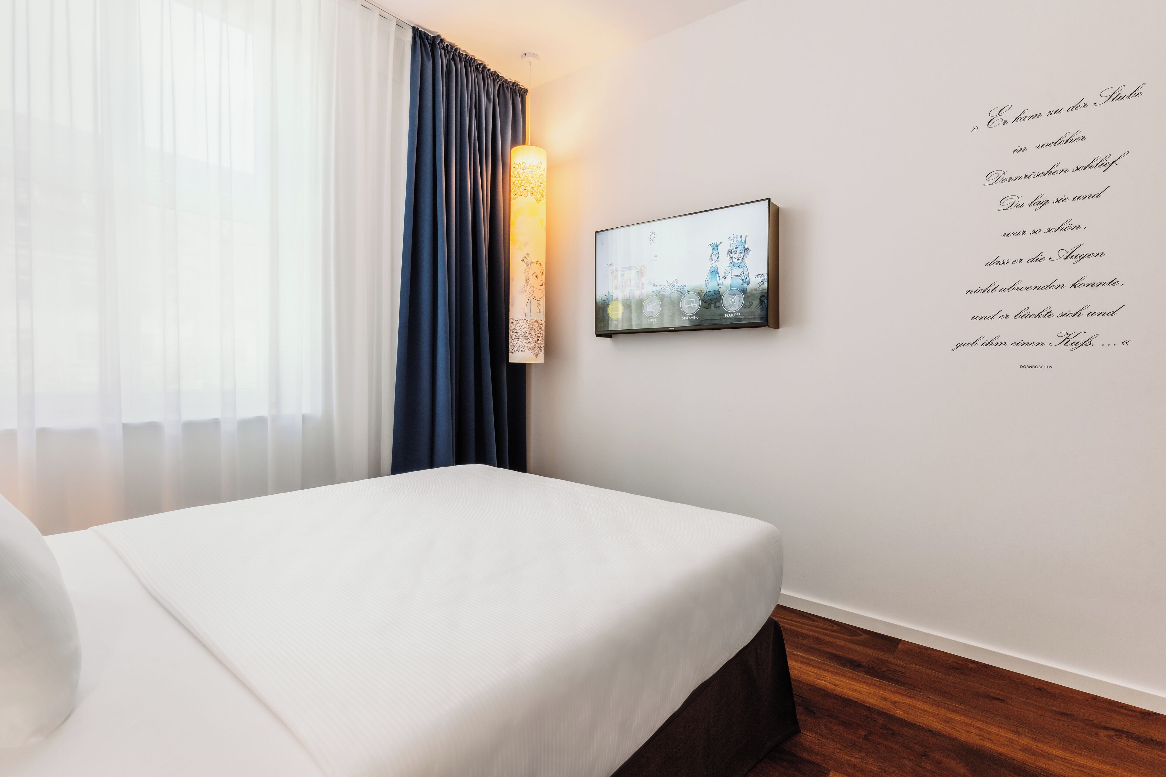 Ein ordentlich bezogenes Bett in einem Hotelzimmer mit einem großen Fenster, einem wandmontierten Fernseher und einem Zitat an der Wand.