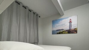 Un dormitorio con una cama, una ventana con cortinas, un cuadro de un faro y una lámpara en el techo.