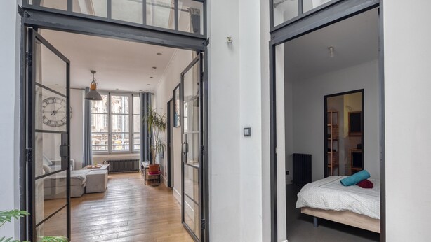 A modern interior with a wooden floor, a bed with a white duvet and blue and red pillows, a large window, and a plant. - Spacious appartment in the heart of Angers - 6 PAX (Angers)