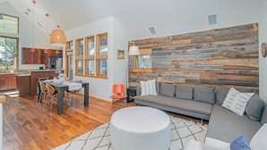 Un salon moderne avec un canapé gris, une table à manger avec des chaises et un coin cuisine doté de meubles en bois et d’une grande fenêtre.