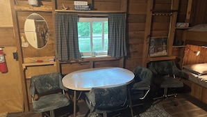 Interior acogedor de una cabaña con una mesa redonda, cuatro sillas, un espejo, una ventana con cortinas, un extintor y una estantería con libros y un letrero.