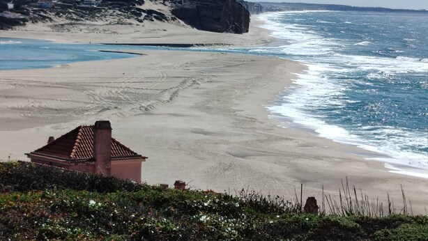 Et kystlandskab med en sandstrand, klippefremspring og en lille bygning med tegltag.