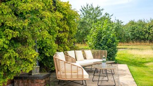 A patio with wicker furniture, including a sofa and a table, set against a backdrop of lush greenery and a brick wall. - Cutbush Farmhouse, Hingham, Norfolk (Hingham)