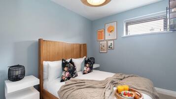 Un dormitorio con cabecera de madera, ropa de cama blanca, almohadas con estampado floral, una mesita de noche con lámpara negra y una bandeja de desayuno sobre la cama.