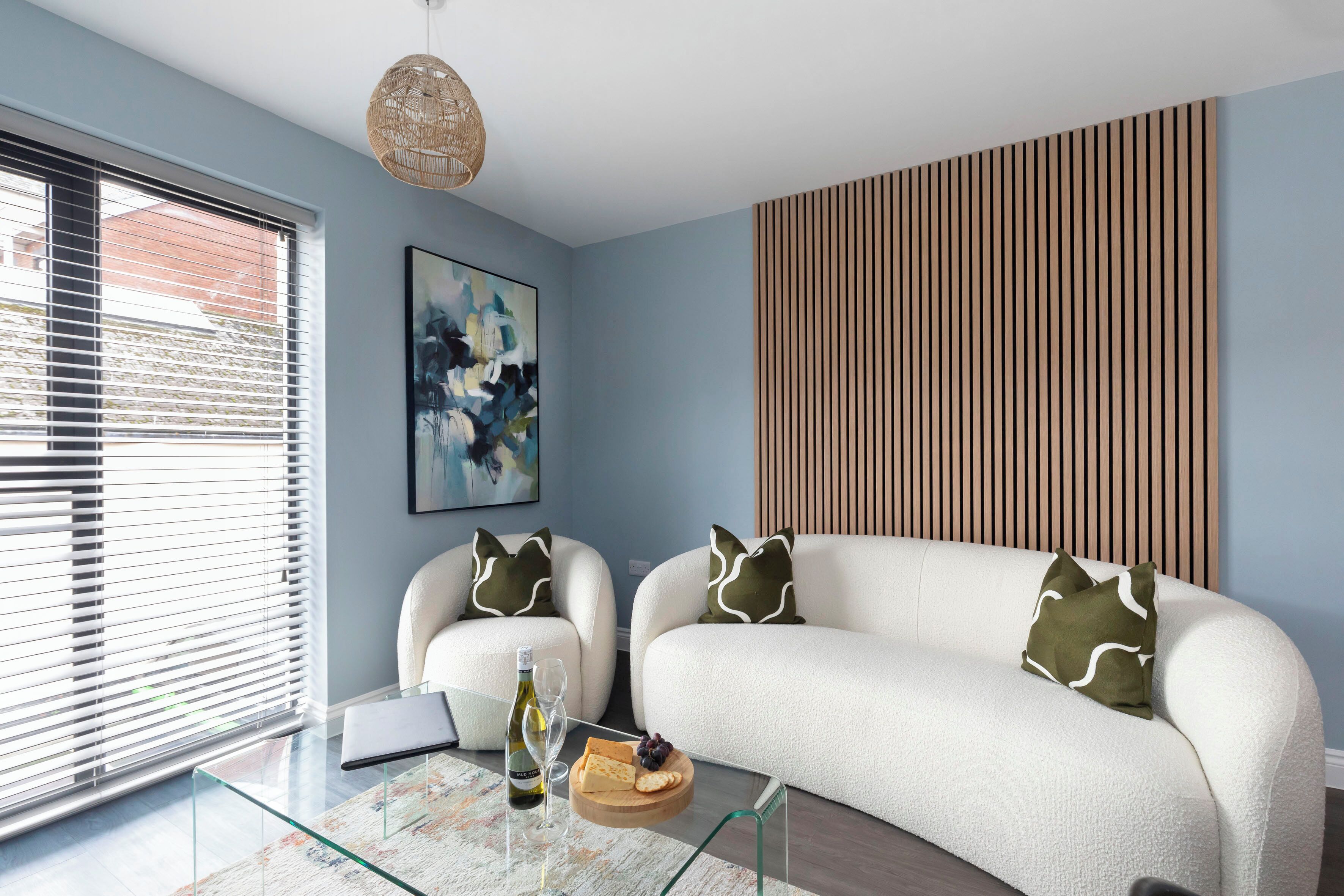 A modern living room with a sofa, two armchairs, a glass coffee table, a painting, and a wooden panel wall.