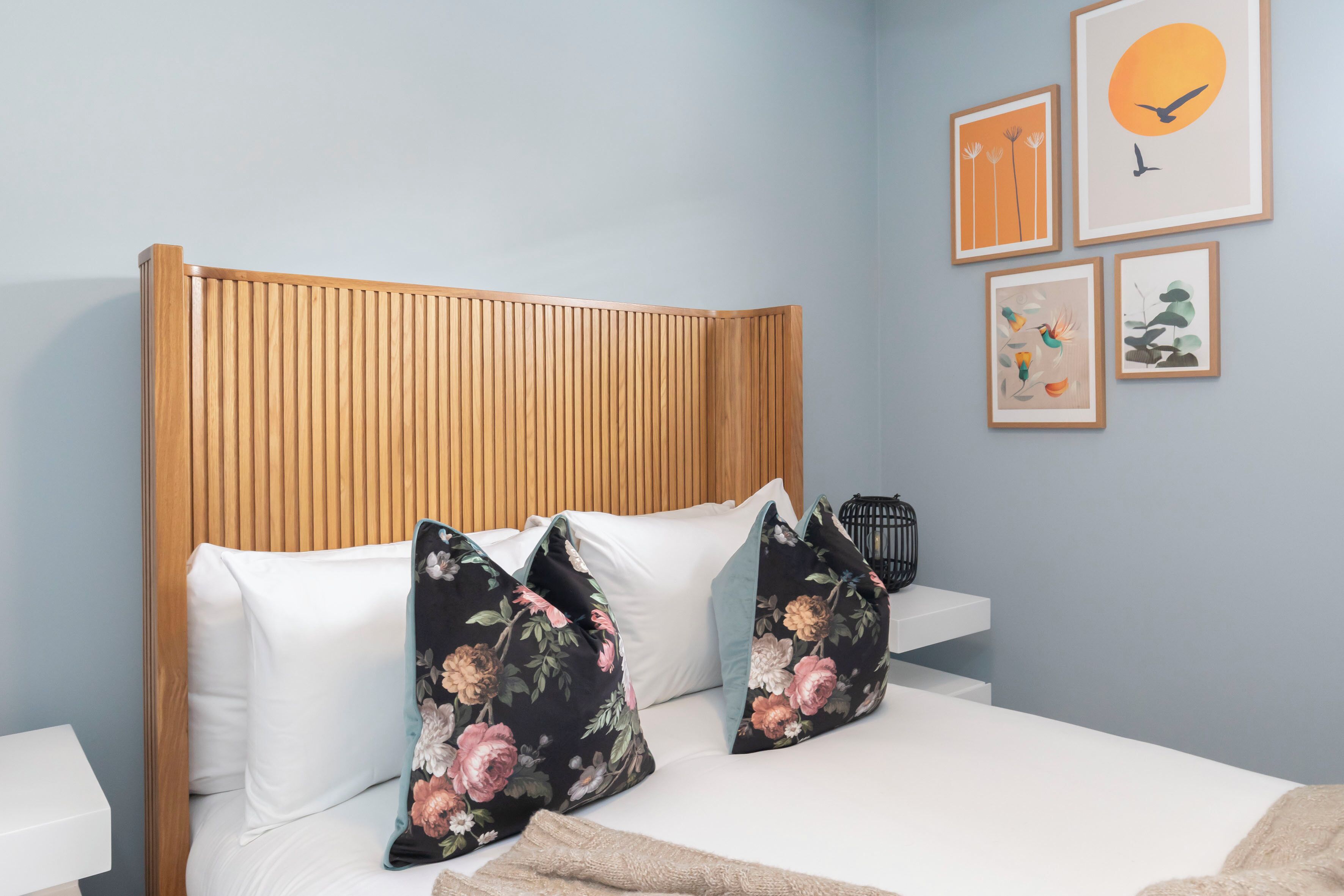 A neatly made bed with white linens and floral pillows, a wooden headboard, and a bedside table with a black object on it.