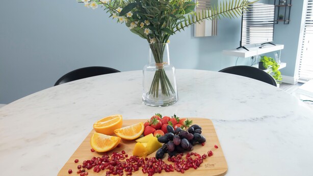 A round marble table with a fruit platter, a vase with flowers, and a modern kitchen in the background. - Beautiful 2 Bedroom Apartment With Parking in The Docks (Gloucester)