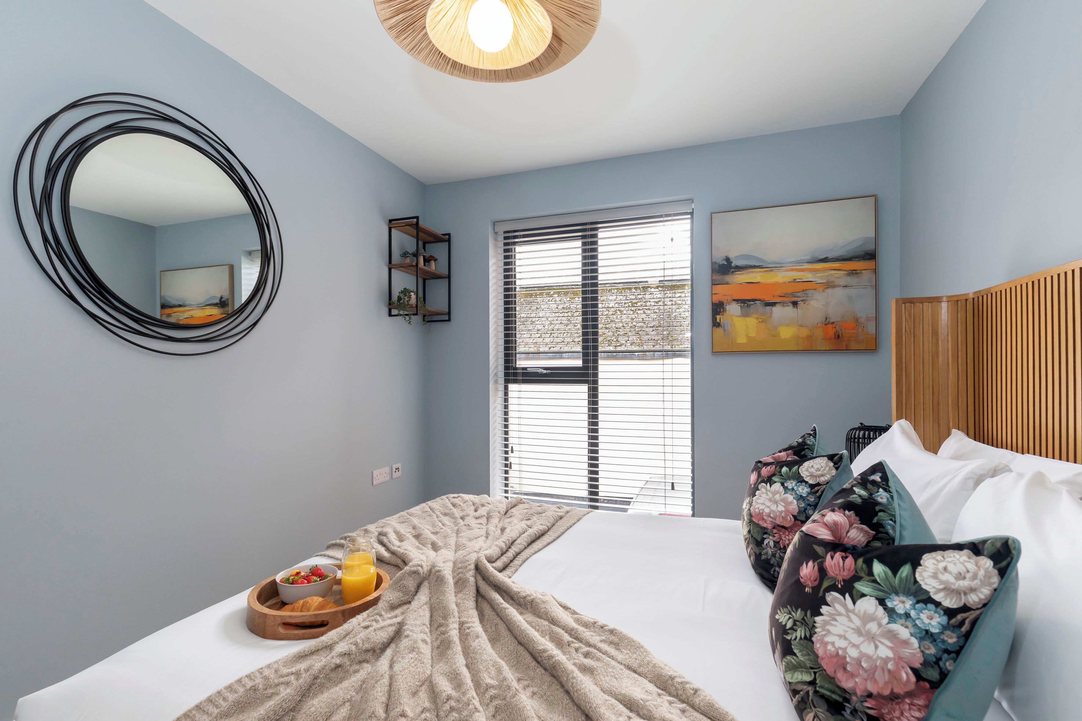 A bedroom with a bed, a round mirror, a window with blinds, a painting, and a wooden headboard.