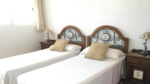 Habitación de hotel con dos camas individuales, cada una con cabecero de madera y colcha blanca. Hay dos mesitas de noche con lámparas y un pequeño adorno en cada una. La habitación tiene un diseño sencillo y limpio, con una paleta de colores neutros.