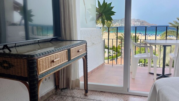 Un balcón con vistas a la playa y al océano, un televisor sobre una mesita consola, y una silla blanca.