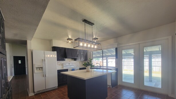 Una cocina moderna con una isla central, armarios oscuros, un refrigerador blanco y un amplio ventanal que proporciona luz natural.