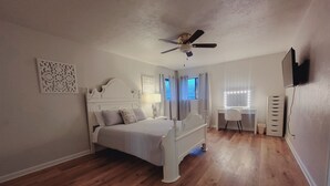 Un dormitorio con cama, escritorio, silla, ventilador de techo y televisor montado en la pared.