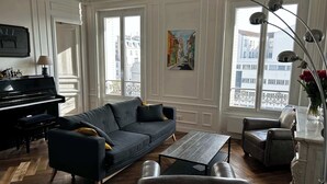 Un salon comprenant un piano, un canapé, une table basse et un fauteuil. De grandes fenêtres offrent une vue sur les bâtiments environnants.