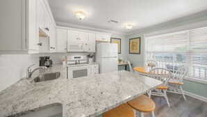 A kitchen with white cabinets, a granite countertop, a sink, and a dining area with white chairs. - Romantic Island Getaway! Just Steps from the Beach (Bradenton Beach)
