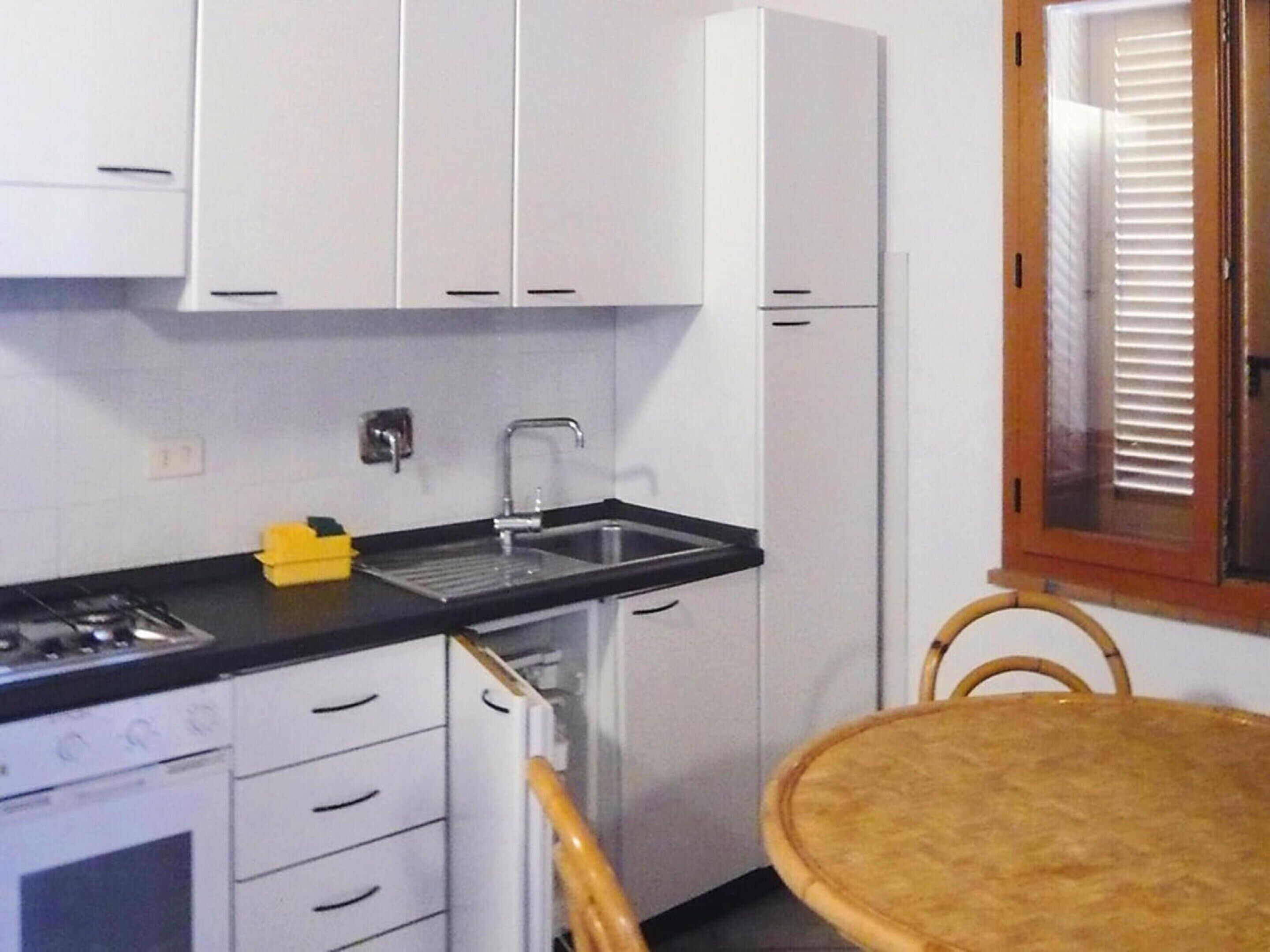 Eine kleine Küche mit weißen Schränken, einer schwarzen Arbeitsplatte, einem Edelstahlspülbecken und einem kleinen Herd. Ein Korbstuhl und ein Fenster mit Jalousien sind ebenfalls vorhanden.