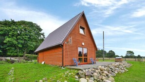 Une cabane en bois avec un toit en pente, une véranda à l’avant dotée de deux chaises et un mur de pierres à l’avant.