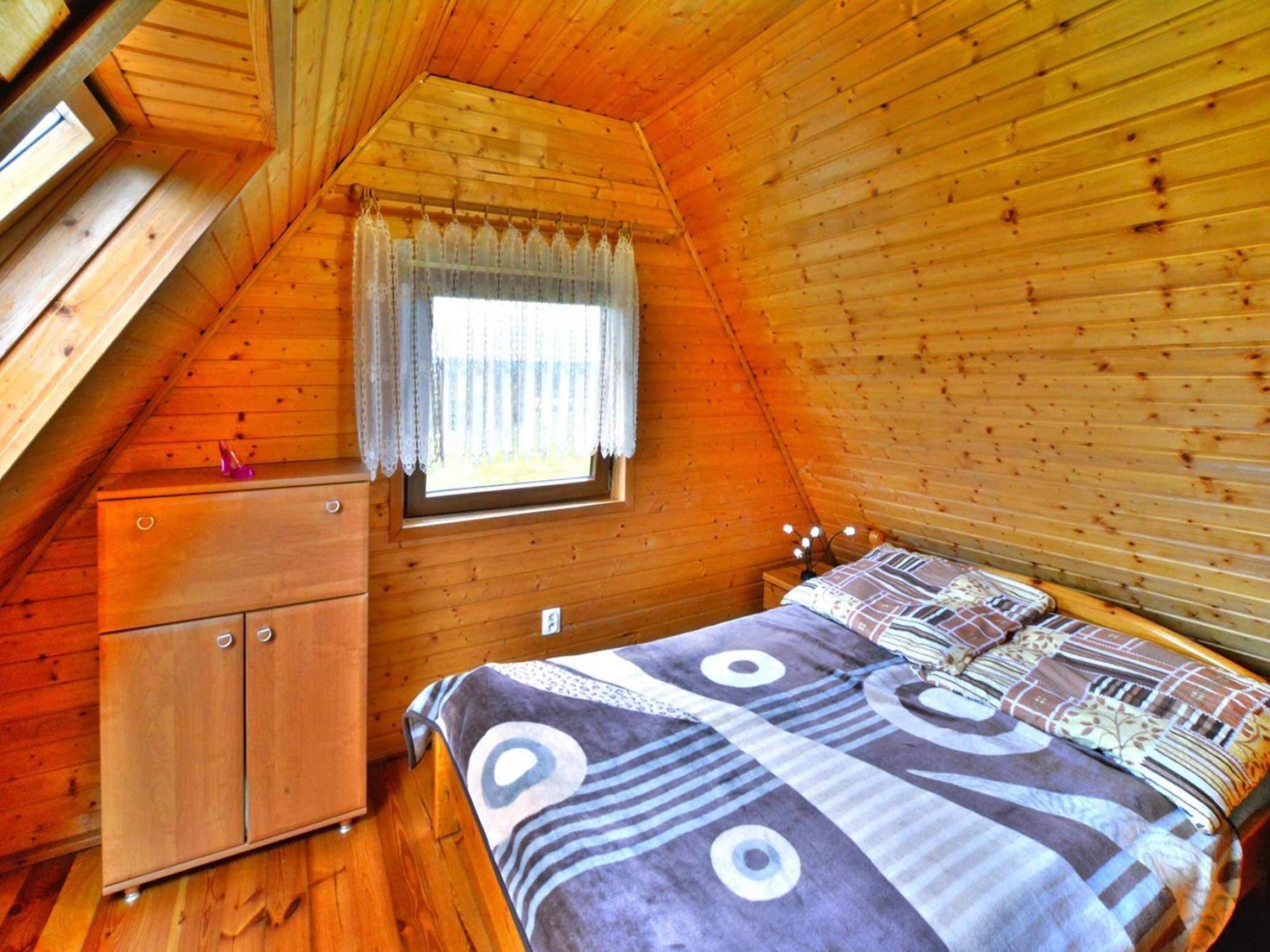 A cozy attic bedroom with a wooden sloped ceiling, a window with a curtain, a bed with patterned bedding, and a small wooden dresser.