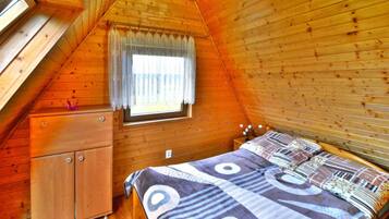 A cozy attic bedroom with a wooden sloped ceiling, a window with a curtain, a bed with patterned bedding, and a small wooden dresser.