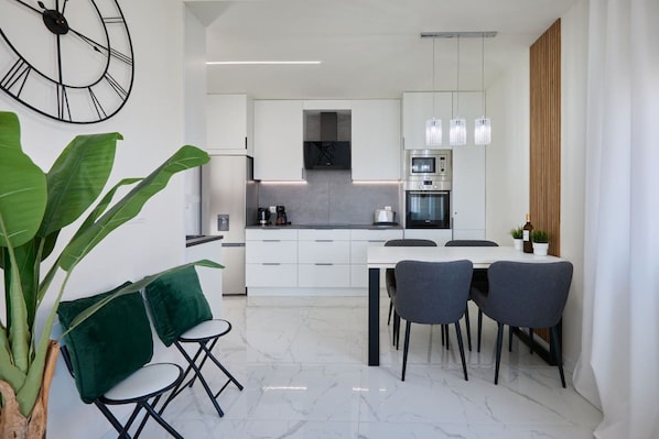 A modern kitchen with white cabinets, a dining table with chairs, and a large green plant. - Casa Loresa Silver (Orihuela)