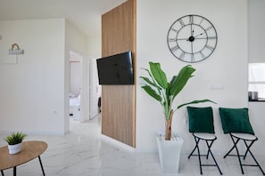 A modern living room with a flat-screen TV, a large wall clock, a potted plant, two green cushions, and a small round table. - Casa Loresa Silver (Orihuela)
