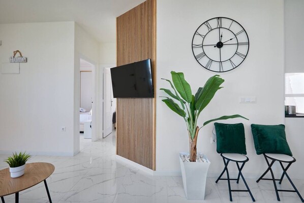 A modern living room with a flat-screen TV, a large wall clock, a potted plant, two green cushions, and a small round table. - Casa Loresa Silver (Orihuela)