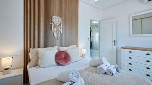 Quarto moderno com parede de ripas de madeira, cama com roupa de cama branca e um sonho apanhador, mesa de cabeceira com abajur e cómoda branca com gavetas.