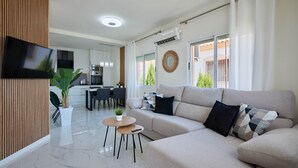 Uma sala de estar moderna com sofá, TV na parede, área de refeições com cadeiras e cozinha ao fundo.