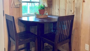 Una habitación con paneles de madera, una mesa redonda y dos sillas, una ventana con cortinas ligeras y una planta en maceta sobre la mesa.