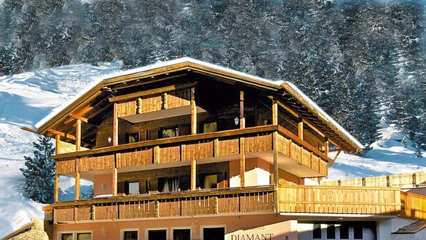 Una cabaña de madera de varios pisos con balcones, identificada con el letrero "DIAMANT", rodeada de montañas nevadas.