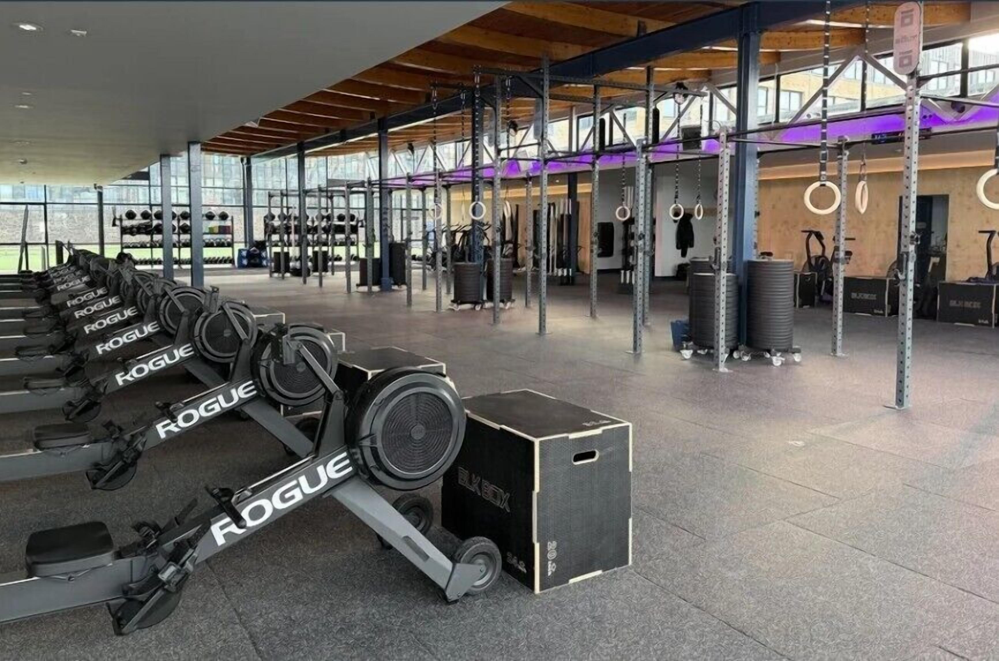 Ein Fitnessstudio mit verschiedenen Trainingsgeräten, darunter ein Rudergerät und ein Ergometer, hohen Decken und großen Fenstern.