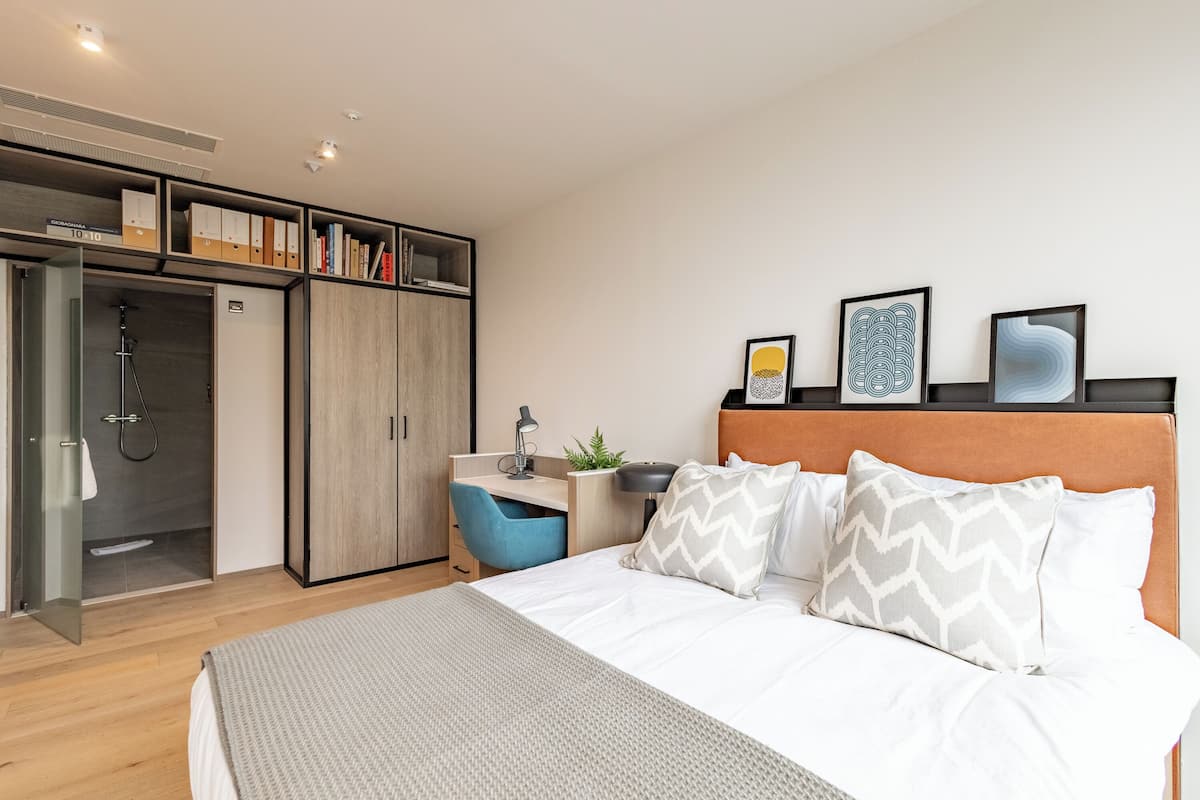 A modern hotel room with a bed, a desk, a shower, and a wardrobe.